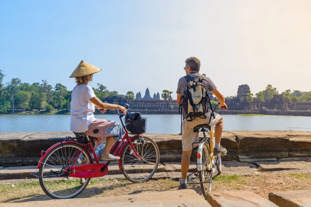 Tourist couple on bicycles admiring the Angkor Wat temple reflected in a water pond.