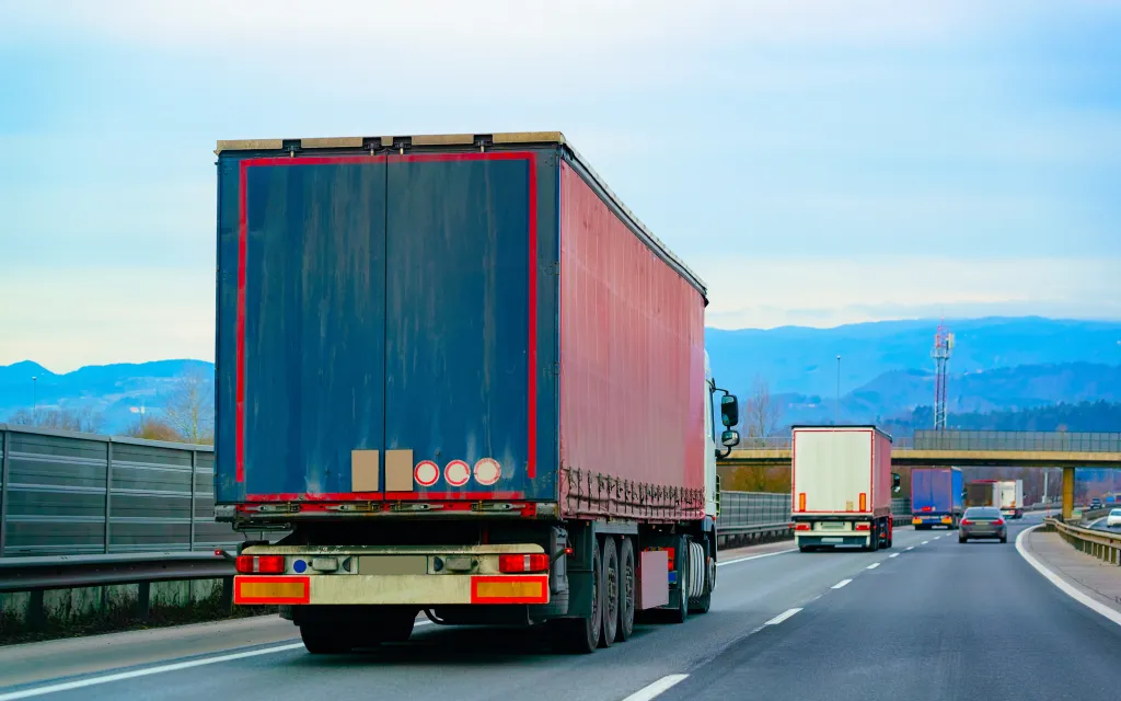 A truck with a blue and red trailer drives on a highway, with other trucks and cars in the distance.