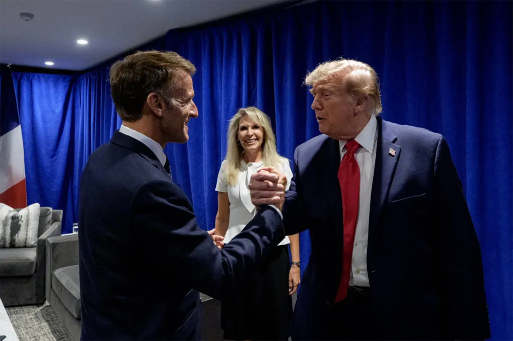 Emmanuel Macron and Donald Trump shake hands, with a woman smiling between them, against a blue curtain backdrop.