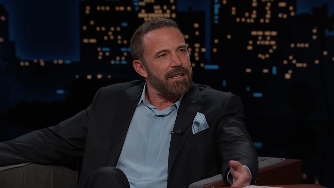 Ben Affleck, wearing a suit and light blue shirt, sits on a talk show set with a city skyline in the background.