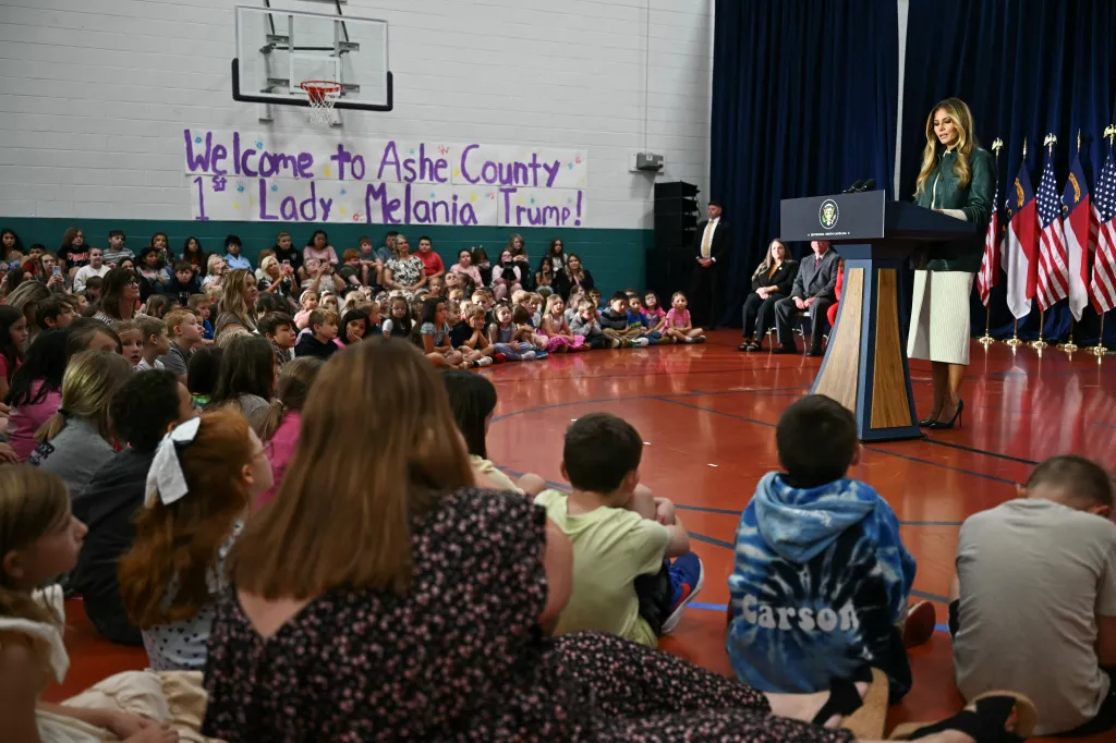Melania Trump speaks at a podium to students sitting on the floor in a school gymnasium.