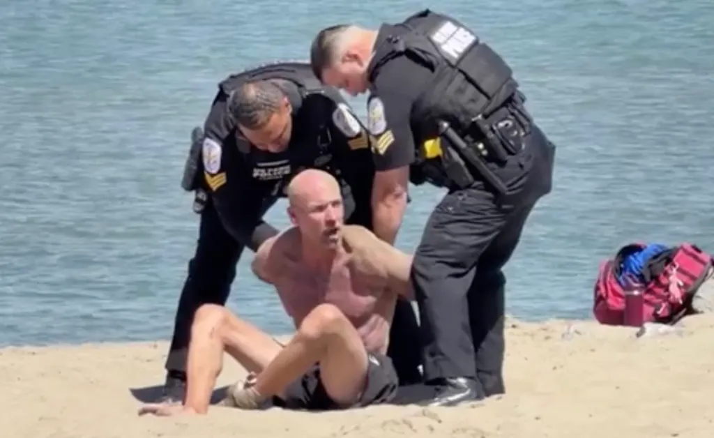 Two police officers apprehending a shirtless man on a beach.