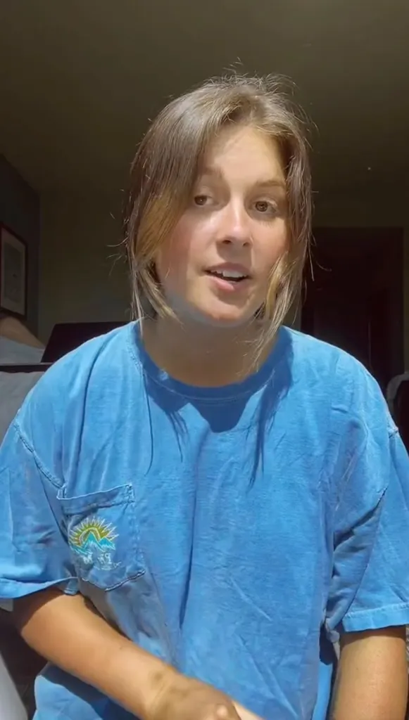 A young woman in a blue t-shirt with a mountain and sun logo on the pocket speaks to the camera.