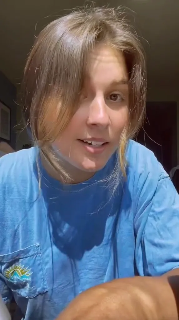A young woman with brown hair smiles, wearing a blue T-shirt with an embroidered patch of mountains and a sun.