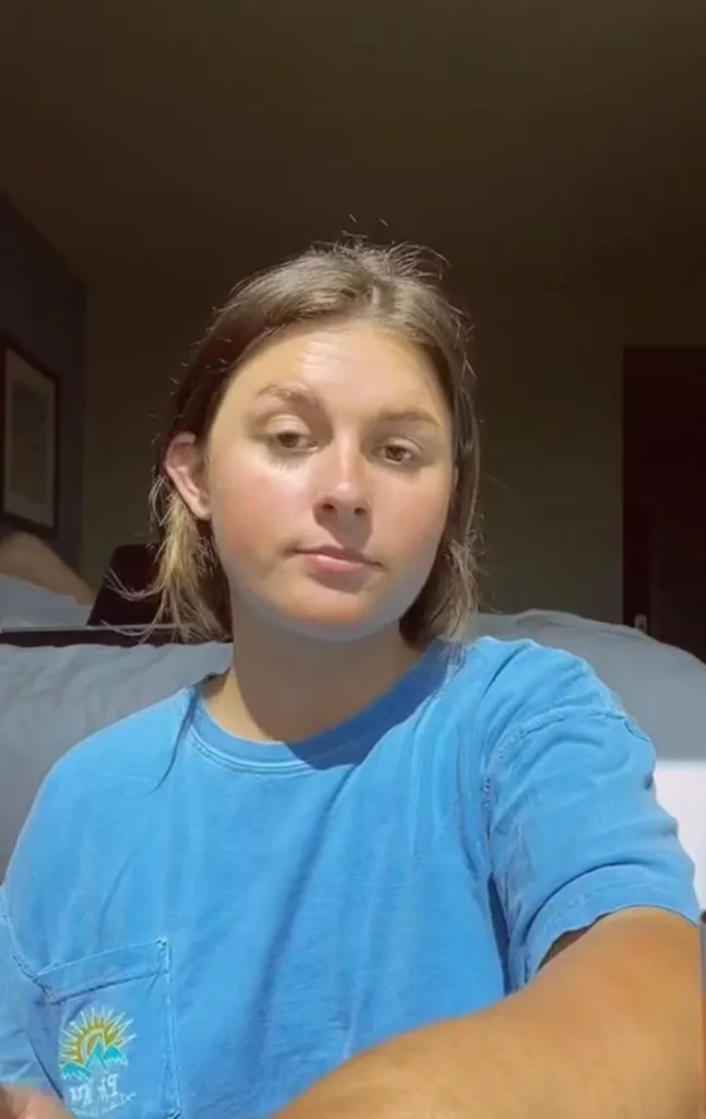 A young woman with light brown hair and a blue t-shirt with a sun graphic on the pocket looks directly at the camera.