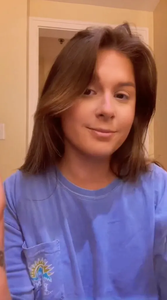 A young woman with brown hair smiles, wearing a blue t-shirt with a pocket featuring a sun and mountains logo.
