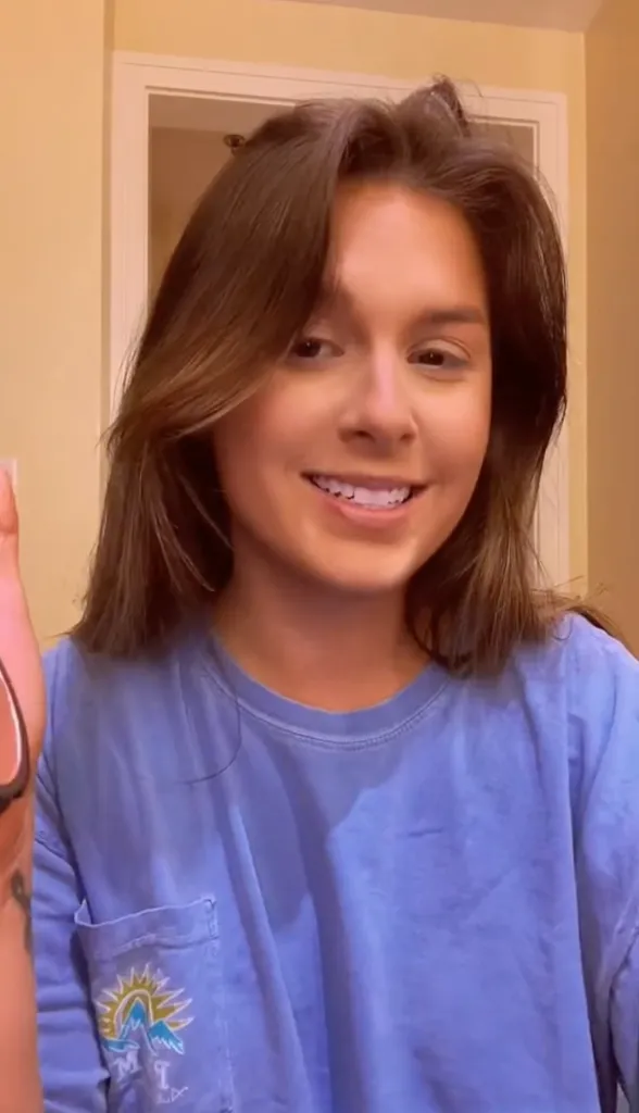 A young woman with brown hair smiles at the camera while wearing a blue long-sleeved shirt with a pocket.