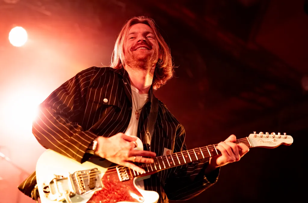 Finneas O'Connell performing live on stage, smiling and playing a white electric guitar, illuminated by a red stage light.