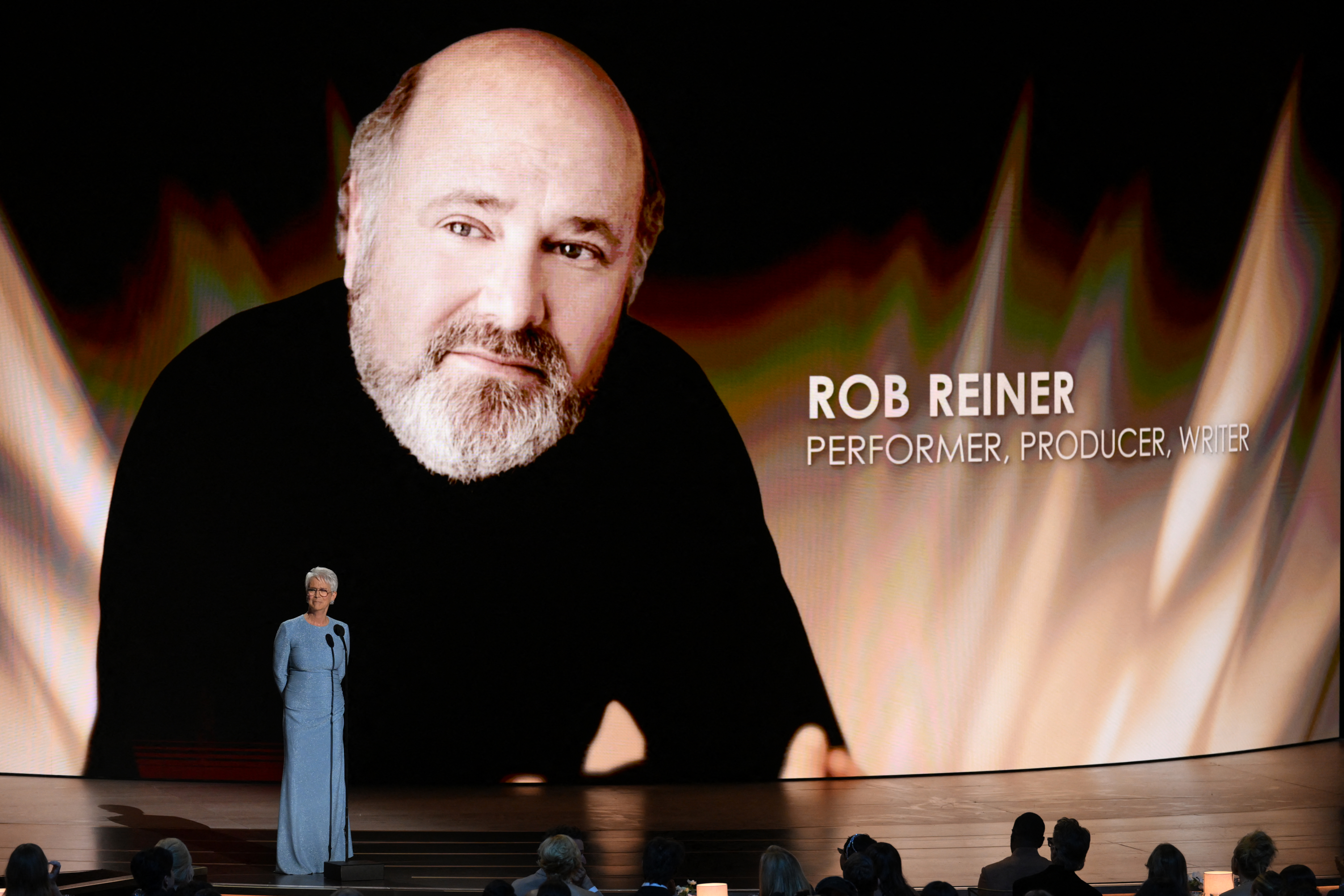 Jamie Lee Curtis on stage in front of a screen displaying a large image of Rob Reiner.