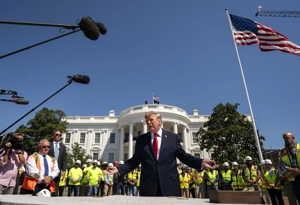President Trump speaks to construction workers near the White House helipad, with the White House in the background.