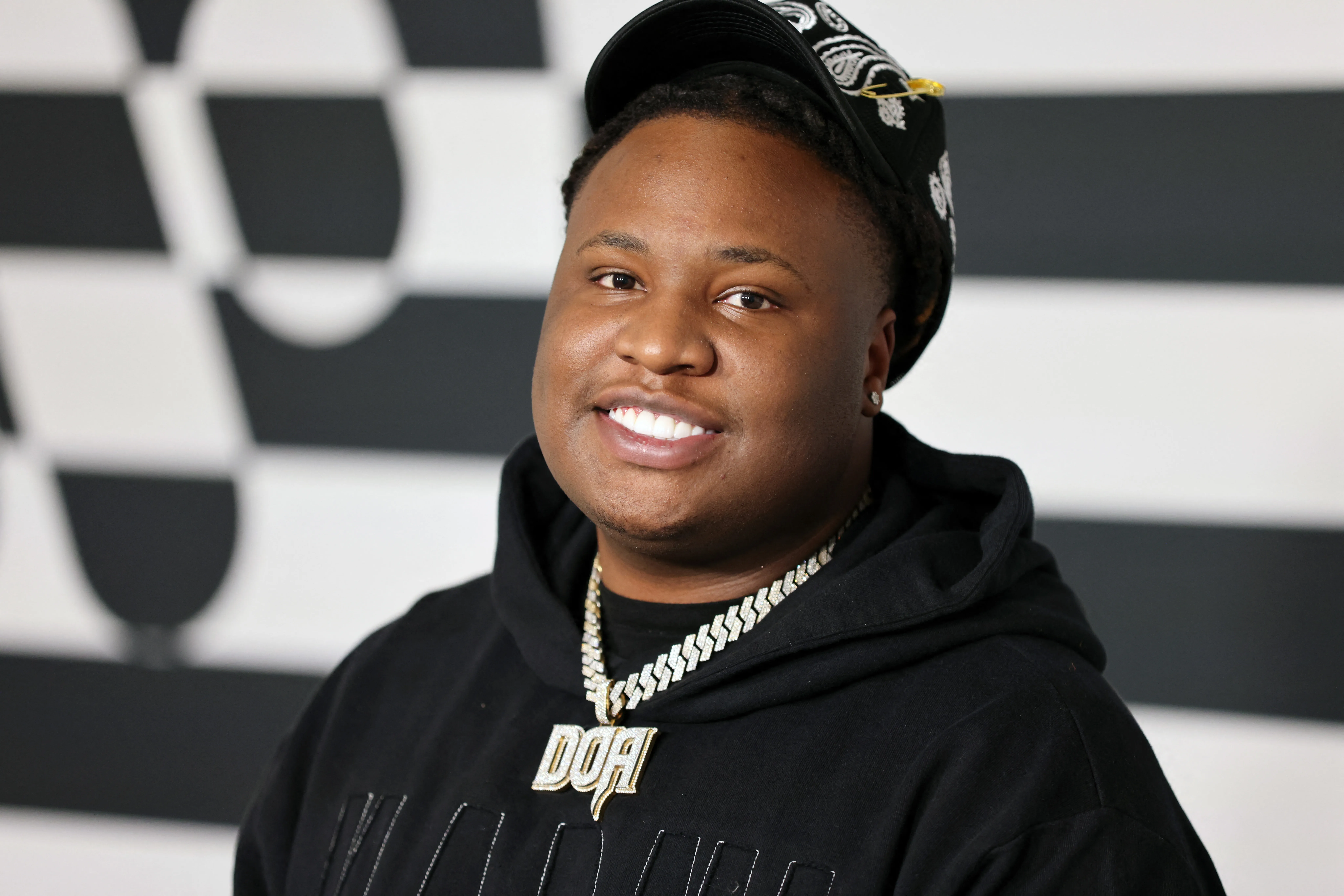 Record producer Tay Keith smiling, wearing a black hoodie and a black cap, with a diamond-encrusted chain that reads 