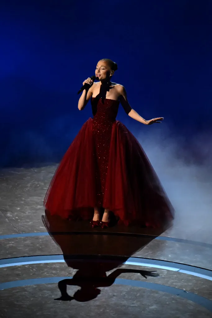 Ariana Grande performing onstage in a red sequined strapless gown with a voluminous tulle overlay, holding a microphone.