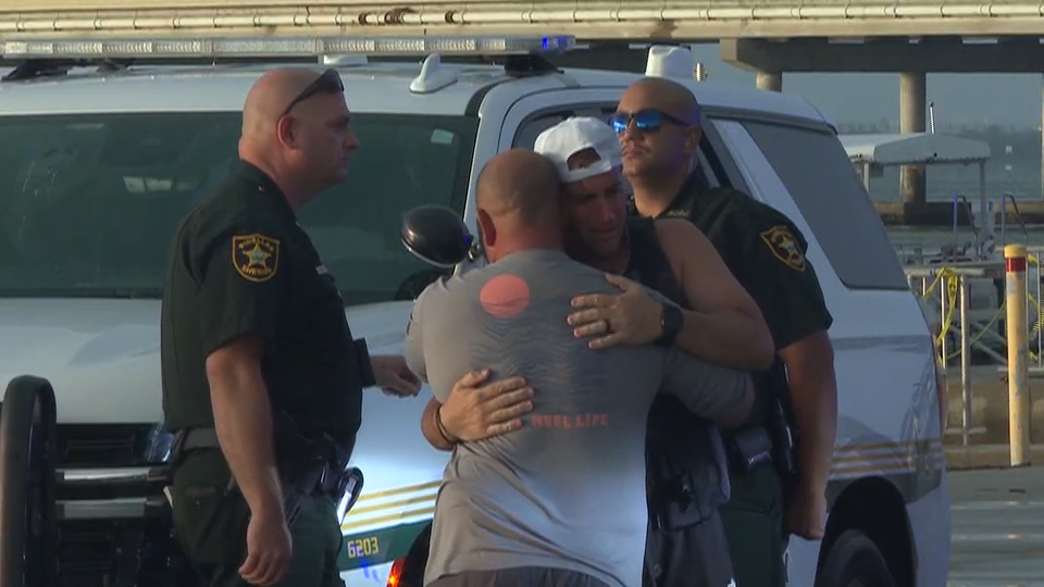 Two men hugging, comforted by two police officers in uniform, near a police vehicle.