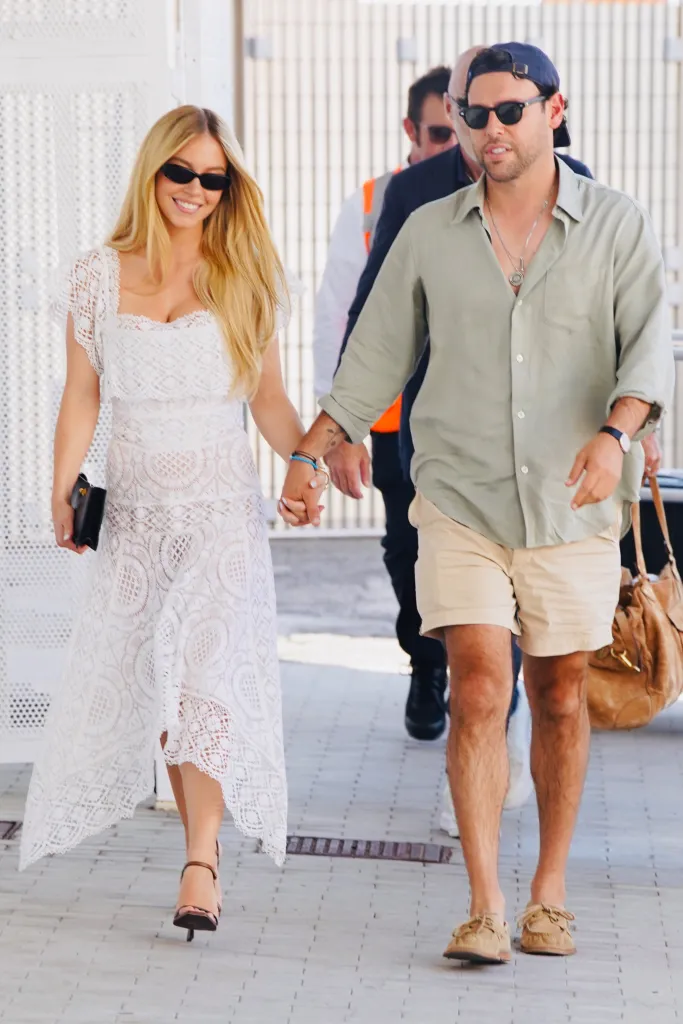 Sydney Sweeney, in sunglasses and a white lacy dress and heels, and Scooter Braun, in sunglasses, a sage green shirt, khaki shorts and a baseball cap, walking hand in hand at Venice Airport.