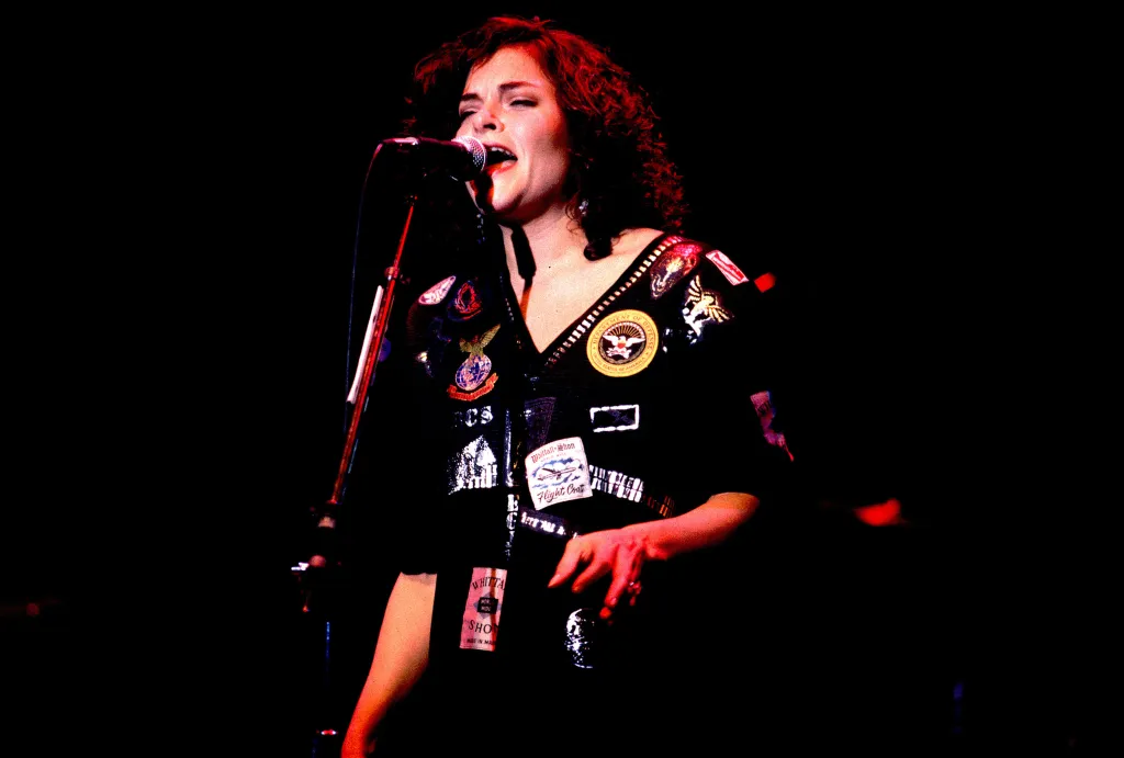 Singer Rosanne Cash performing on stage in a black top adorned with various badges and patches.