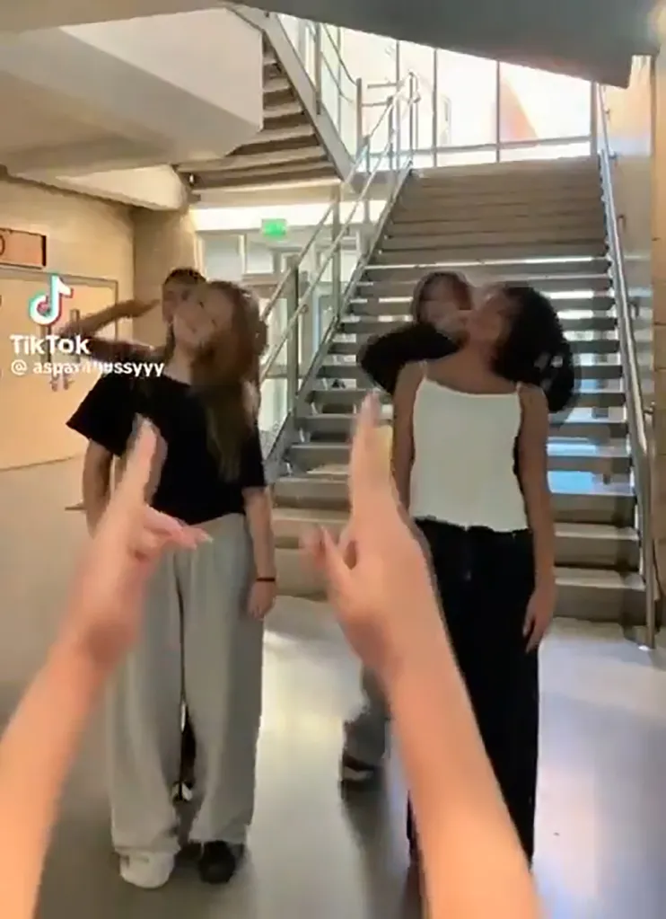 A group of young people gathered in a hallway with a staircase behind them, engaging in a social media trend mocking the death of Charlie Kirk.