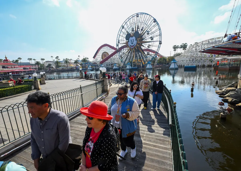 Visitors wait in line on a bridge at Disney California Adventure, with the Pixar Pal-A-Round Ferris wheel in the background.