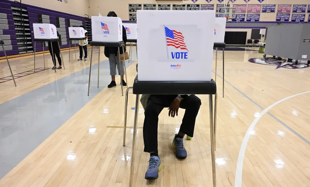 Voters filling out ballots at voting booths in a high school gymnasium.