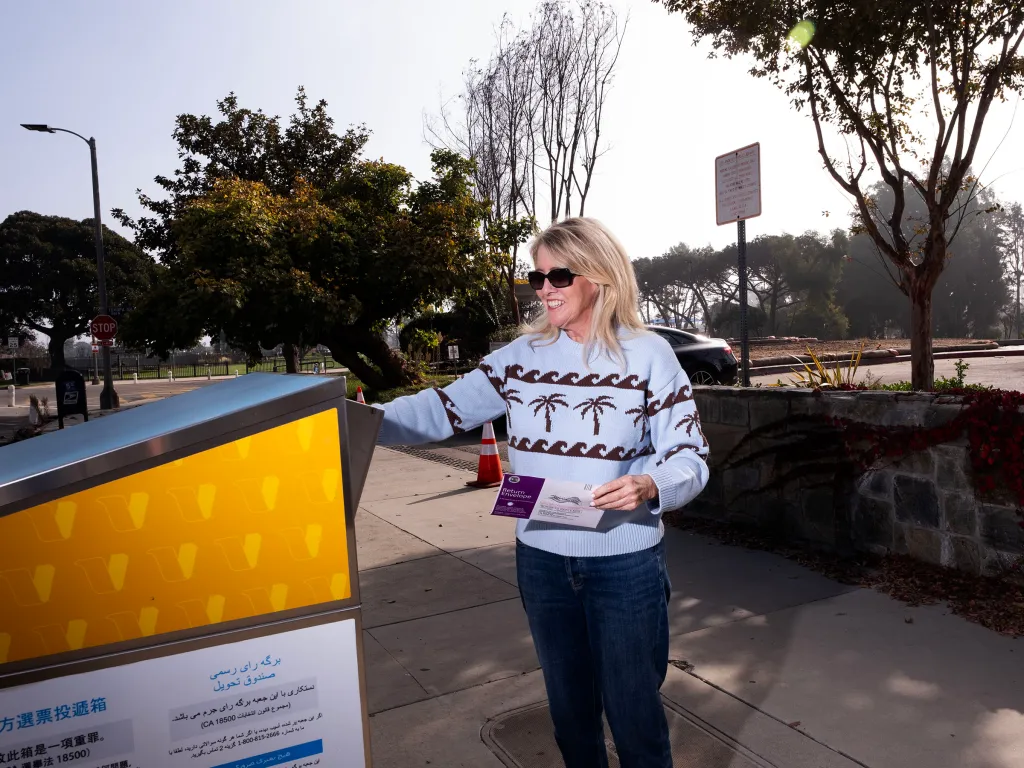 A smiling woman wearing sunglasses and a light blue sweater with palm trees drops a ballot into an official ballot drop box.