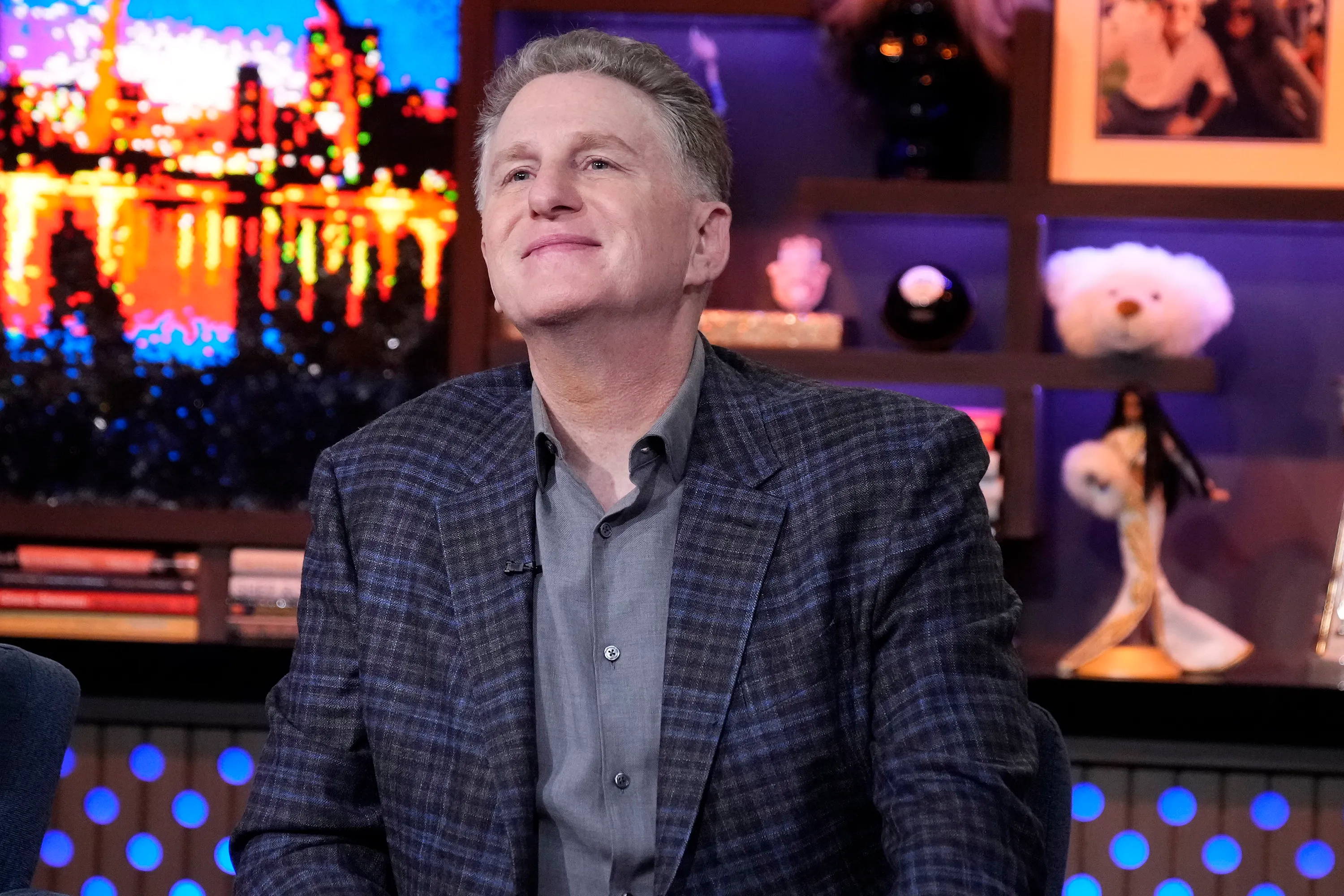 Michael Rapaport smiling while wearing a gray shirt and blue plaid sport coat.
