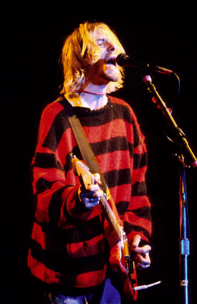 Kurt Cobain performing live onstage, singing into a microphone and playing a red Fender Mustang guitar, wearing a red and black striped sweater.