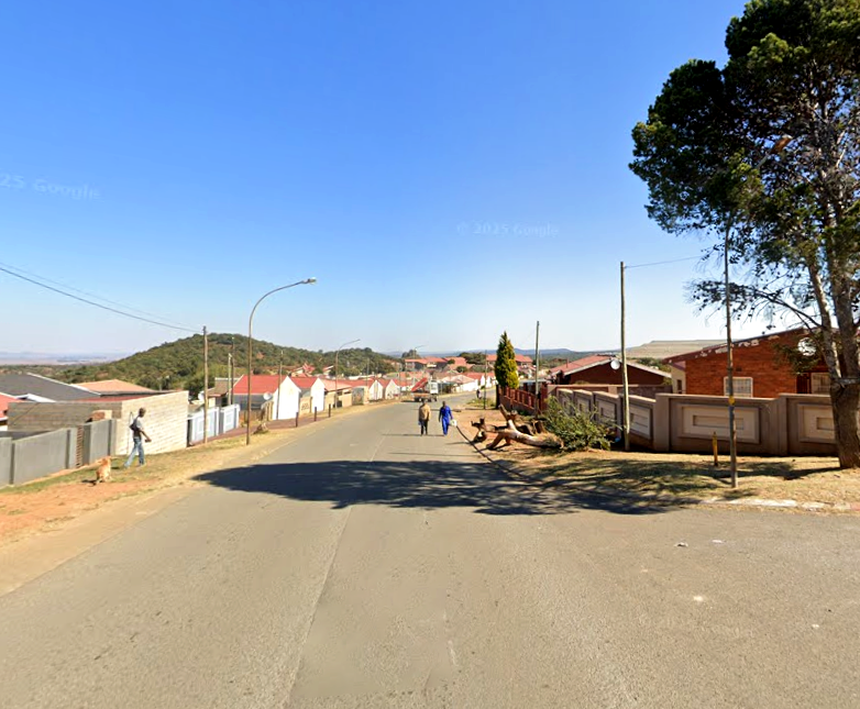 A street in Wedela township, South Africa, with houses, fences, and hills in the background.