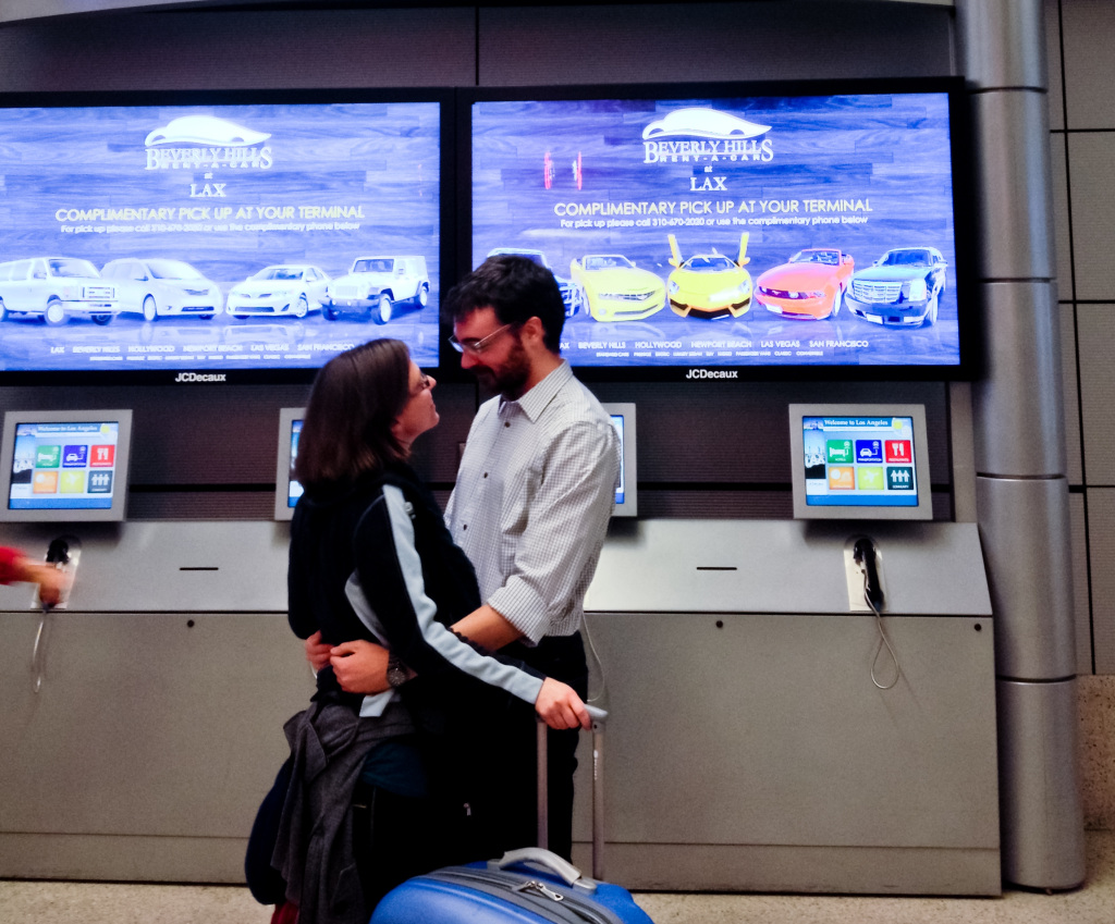 A couple embracing near a Beverly Hills Rent-A-Car advertisement at Los Angeles Airport.
