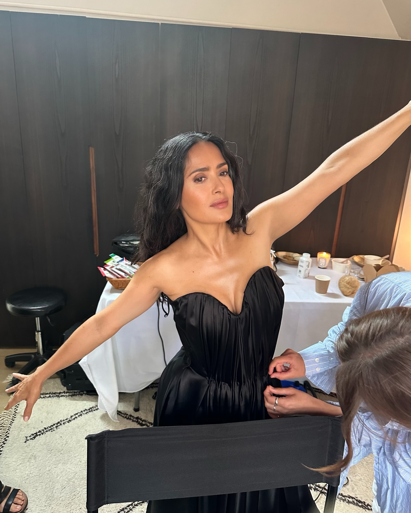 Salma Hayek poses in a black dress while someone adjusts it.