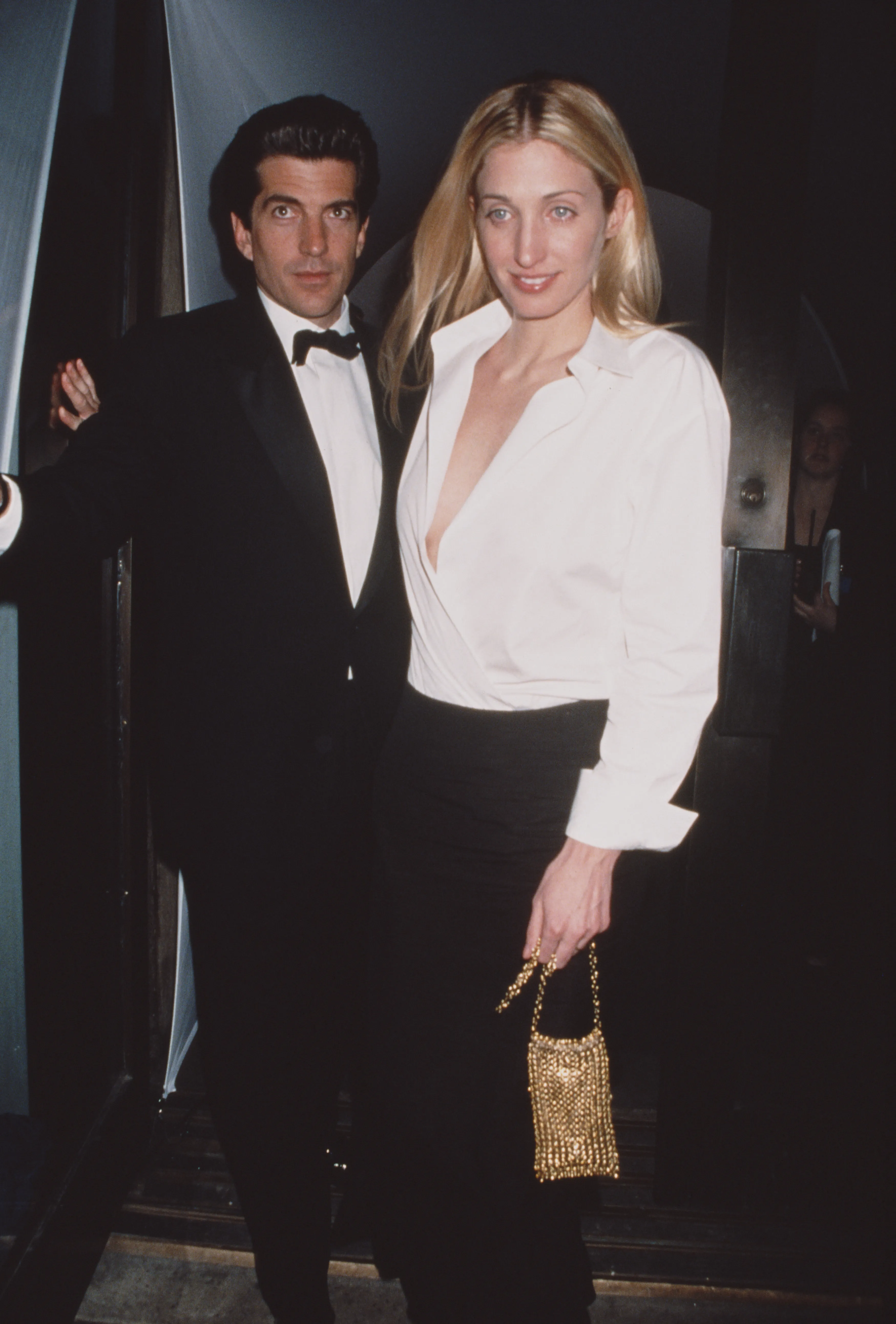 John F. Kennedy Jr. in a tuxedo stands next to Carolyn Bessette-Kennedy in a white shirt and black skirt.