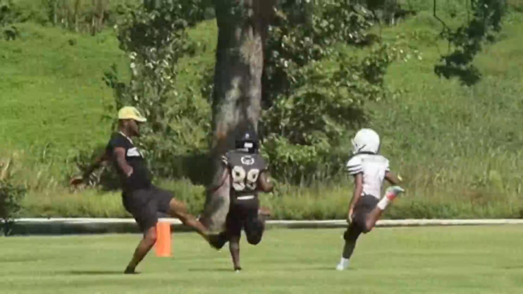 An adult trips a 9-year-old boy in a football uniform on a grassy field.