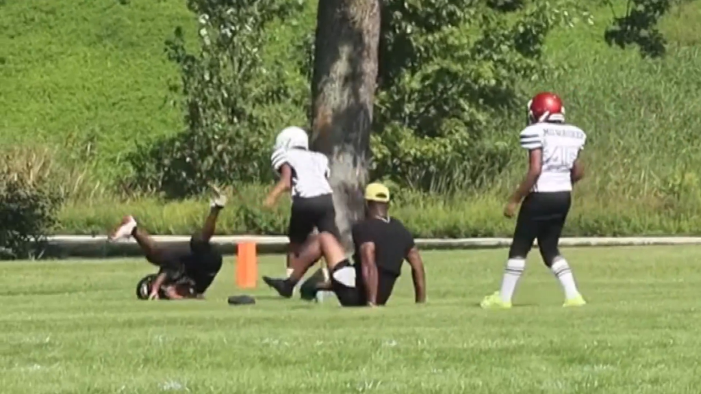 A man in a yellow cap trips a 9-year-old boy in a football uniform during a youth football game.