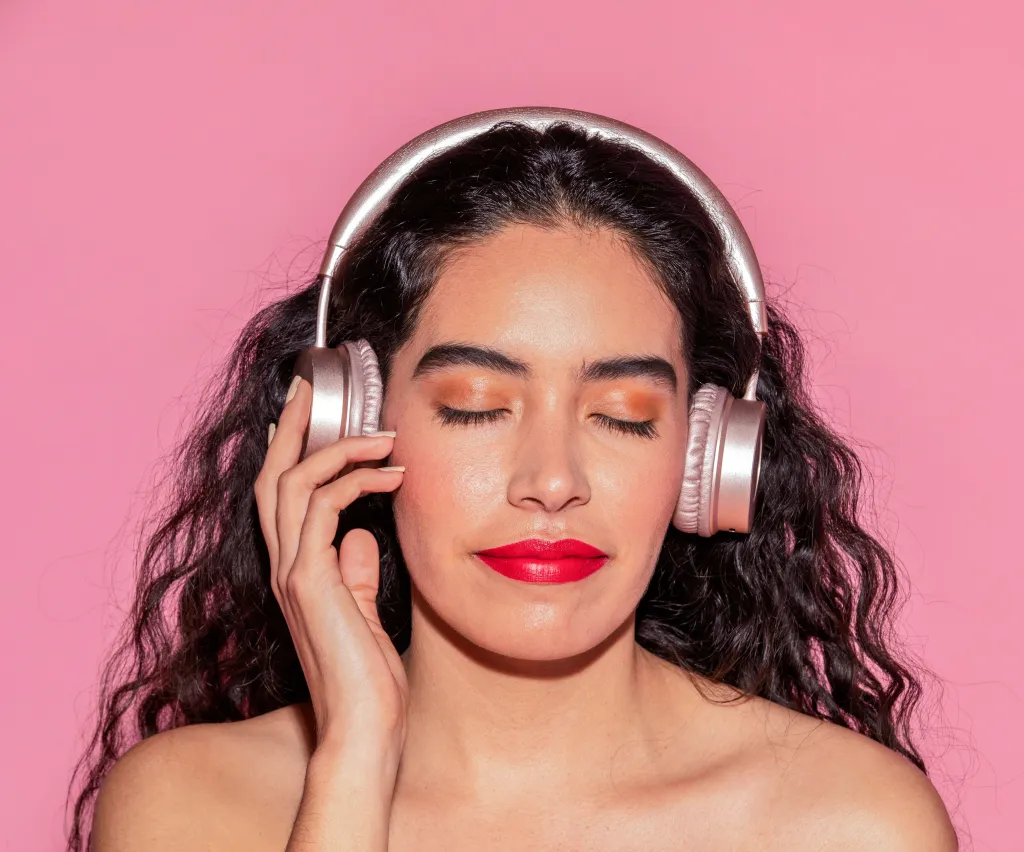 A Hispanic woman with curly hair, red lipstick, and closed eyes enjoys music through headphones against a pink background.