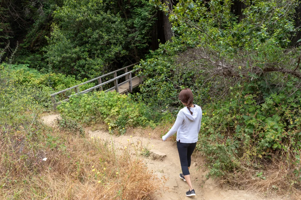 A woman in athletic wear hikes a dirt trail leading to a wooden bridge in a lush green forest in Big Sur, California.