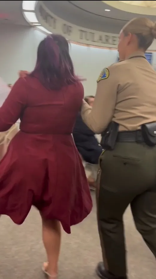 A woman in a red dress is escorted out of a Tulare County Board of Supervisors meeting by a police officer.