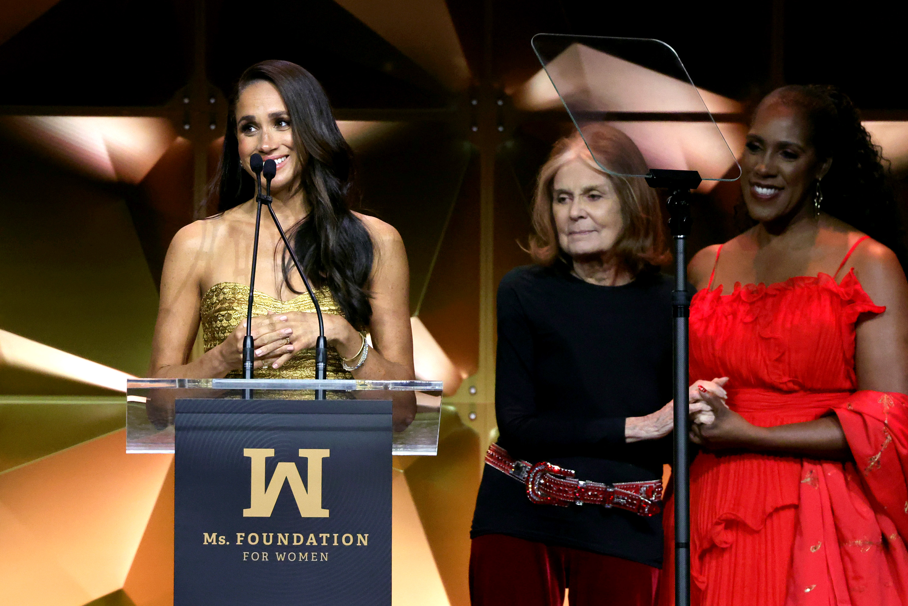 Meghan, Duchess of Sussex, speaking at a podium with Gloria Steinem and Teresa Younger by her side.