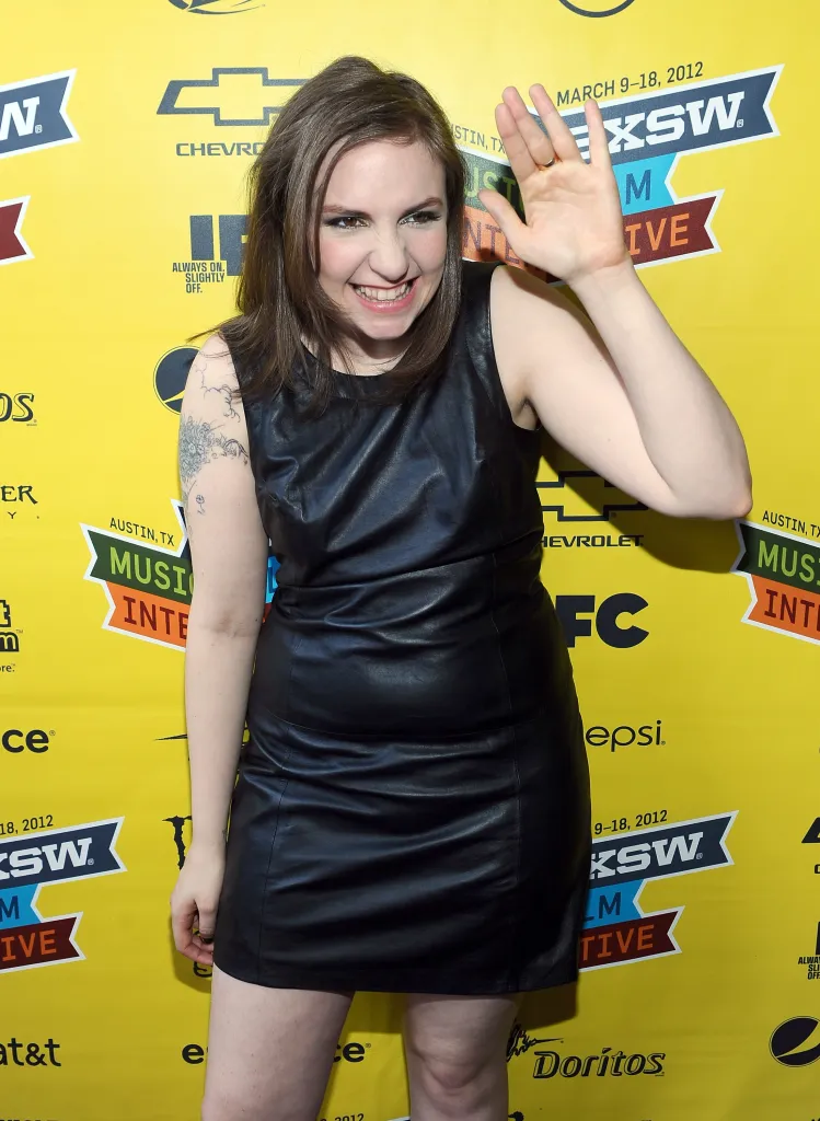 Lena Dunham on the red carpet at the SXSW festival, raising one hand, smiling, and wearing a black leather dress.