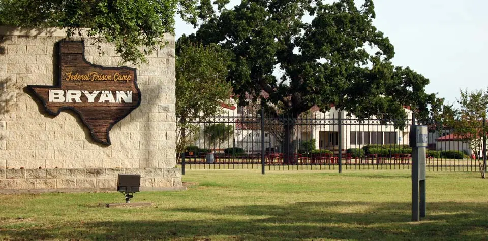Sign for Federal Prison Camp Bryan, with buildings and a fence in the background.