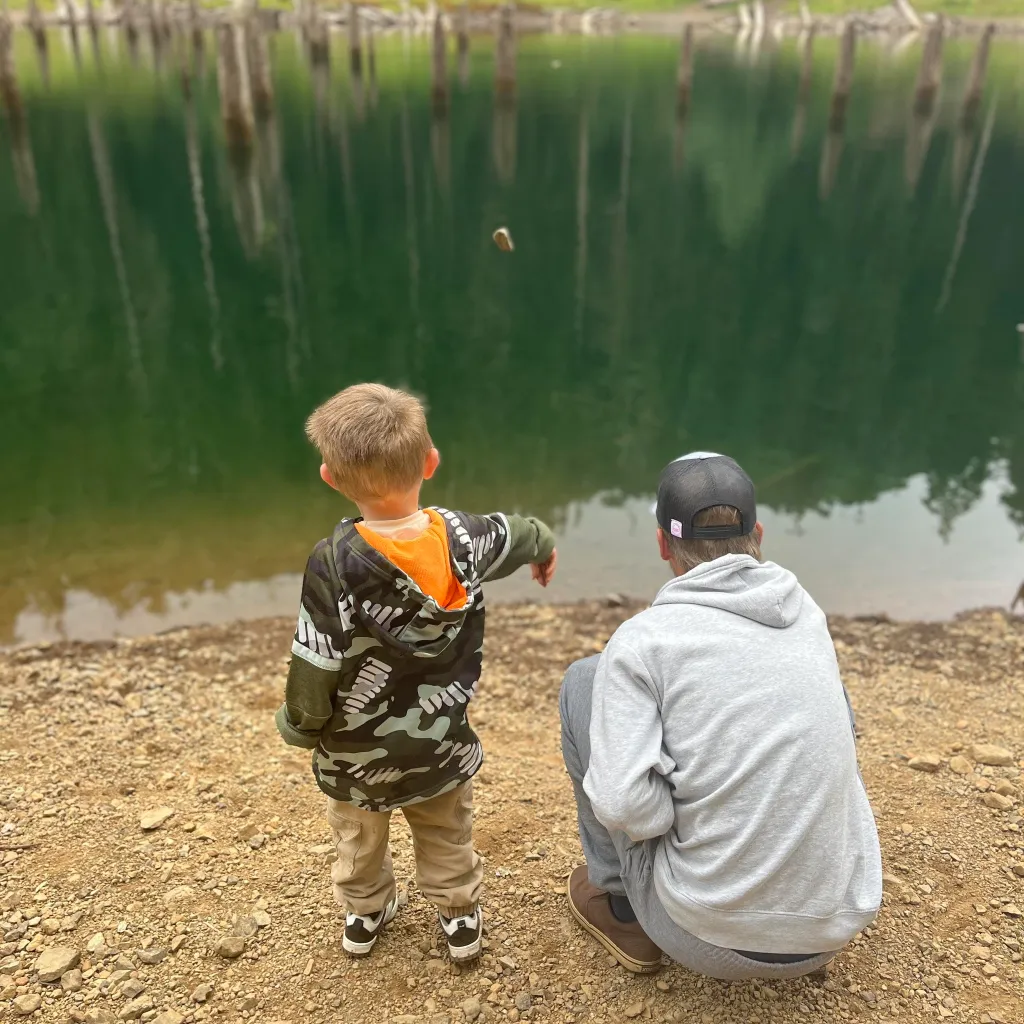 A young child throws a rock into a lake with a person crouching next to them.