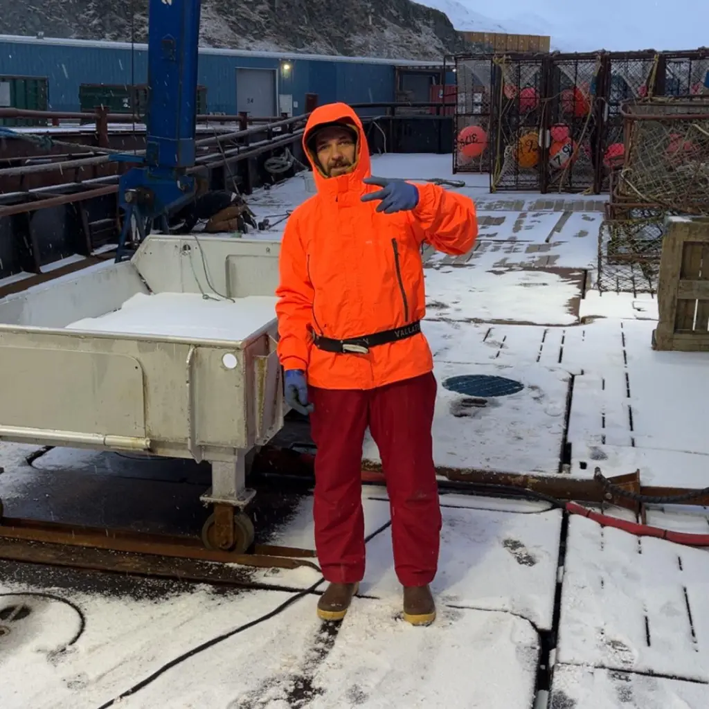 Todd Meadows, from Deadliest Catch, poses in a bright orange jacket and red pants on a snowy boat deck.