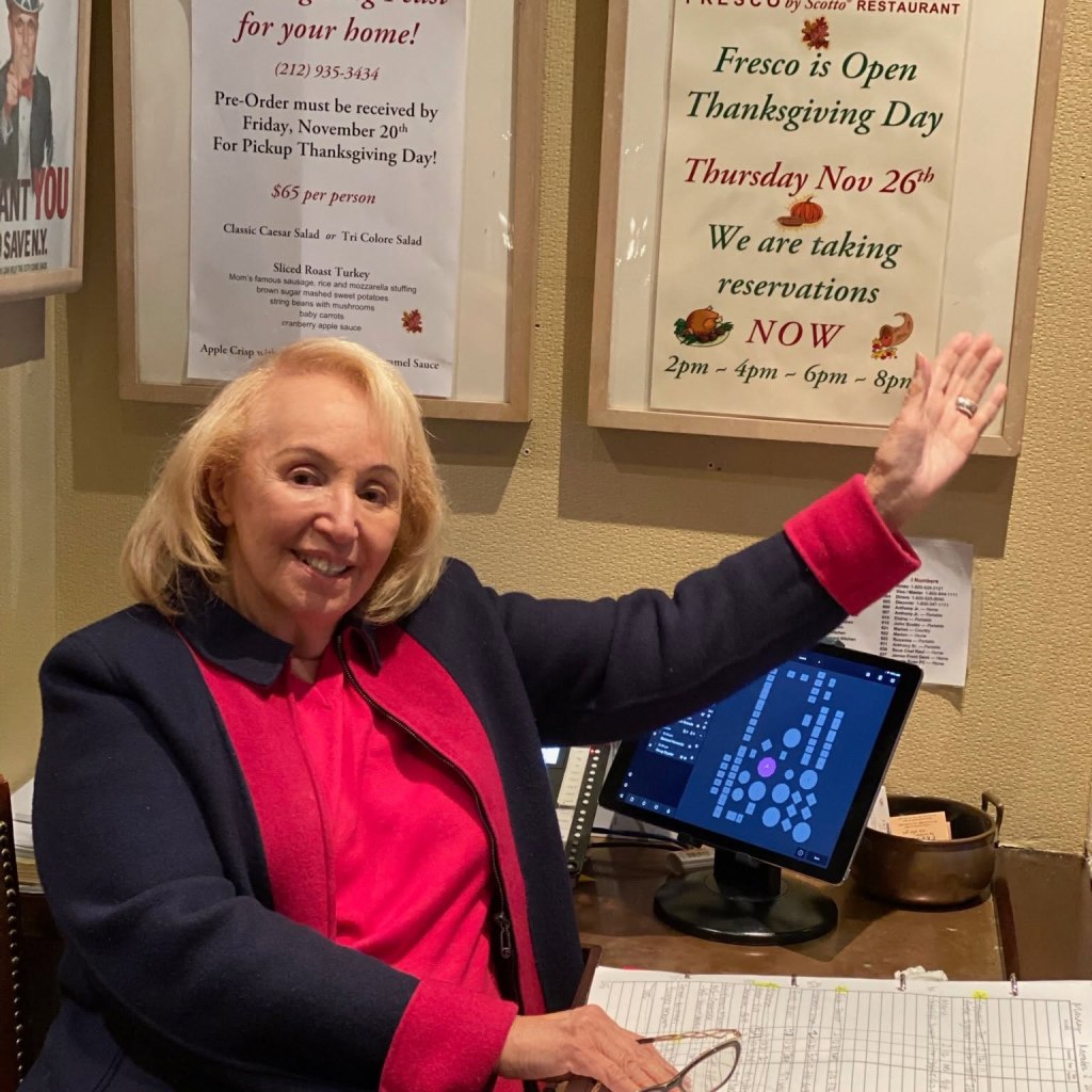 Rosanna Scotto in a restaurant, gesturing towards a sign advertising Thanksgiving Day reservations and pre-orders.