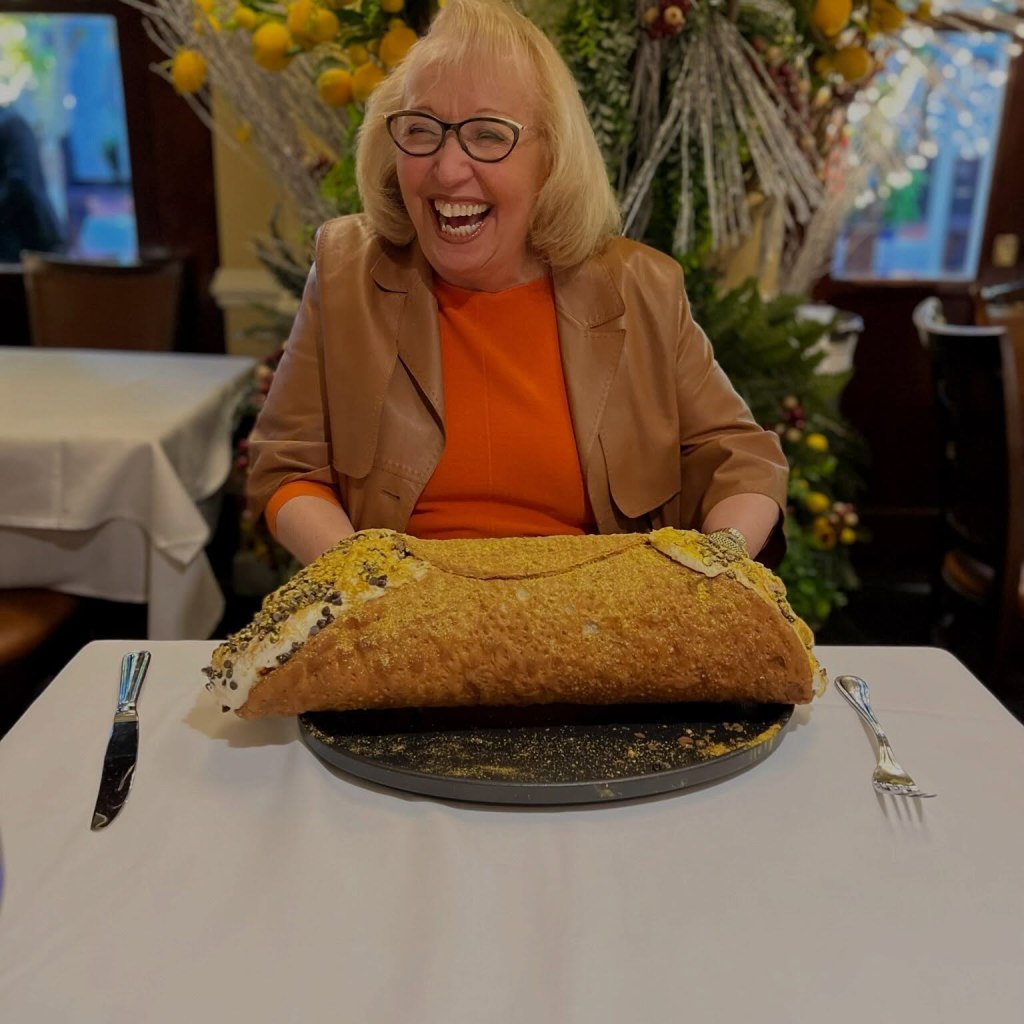 A woman laughs happily behind an enormous cannoli on a table.