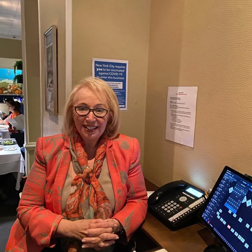 A woman in a coral blazer and patterned scarf smiles in front of a sign stating NYC requires vaccination against COVID-19 to enter.