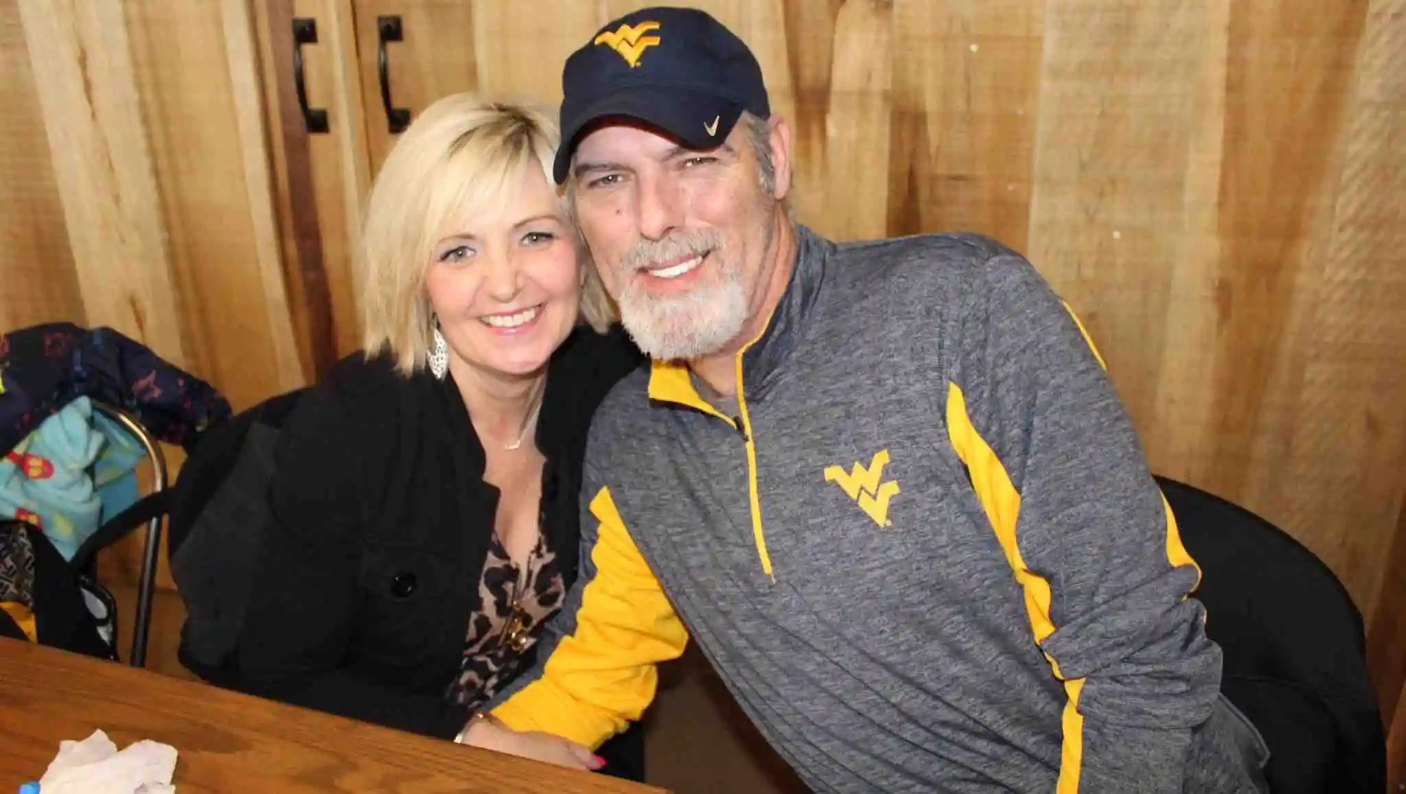 Tina Duckworth, with blonde hair, smiling next to a man wearing a baseball cap and a gray and yellow shirt.