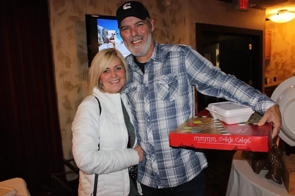 A smiling woman with blonde hair stands next to a smiling man with a beard and a black hat who is holding a
