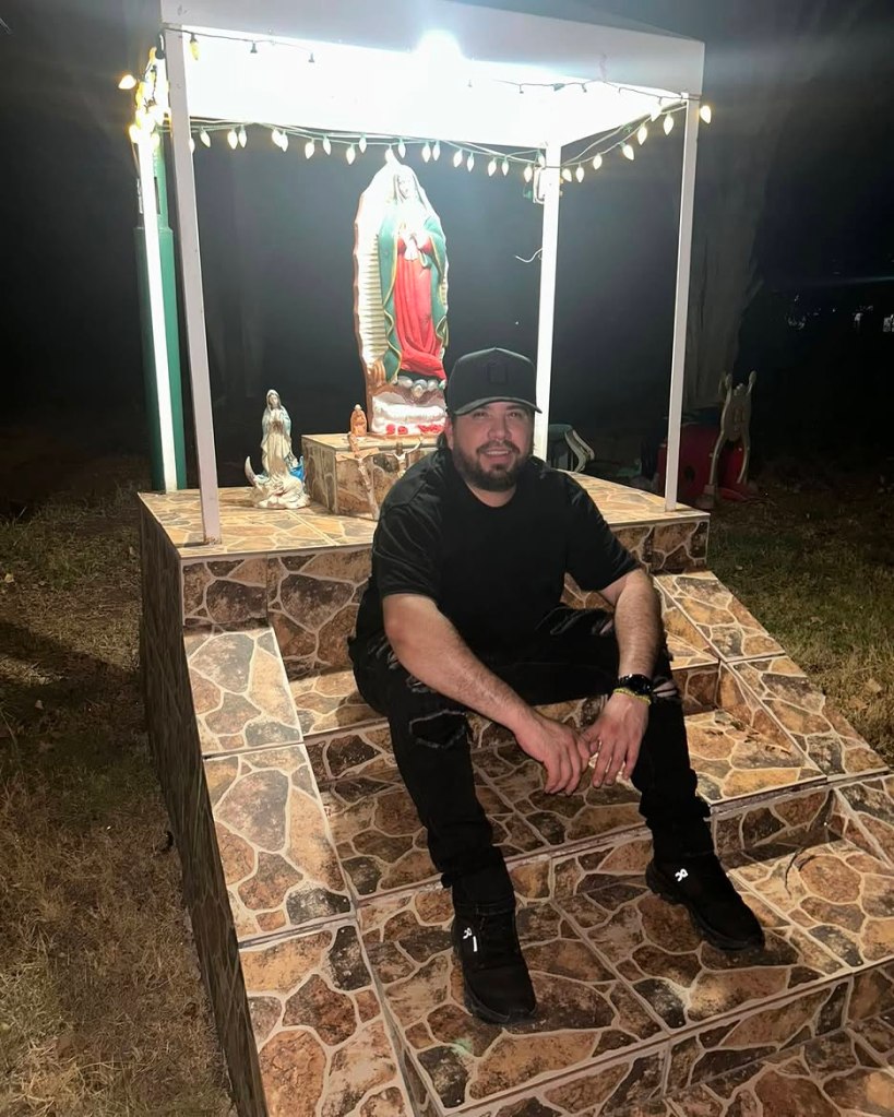 A man in a black shirt, black ripped jeans, and a black baseball cap sits on tiled steps in front of a statue of the Virgin Mary at night.