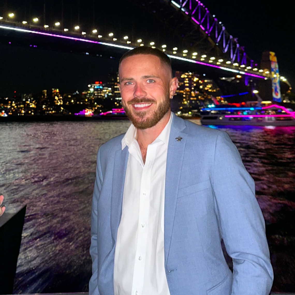 Nathan Gallagher smiling in a light blue suit jacket and white shirt, with a city skyline and bridge lit up at night in the background.