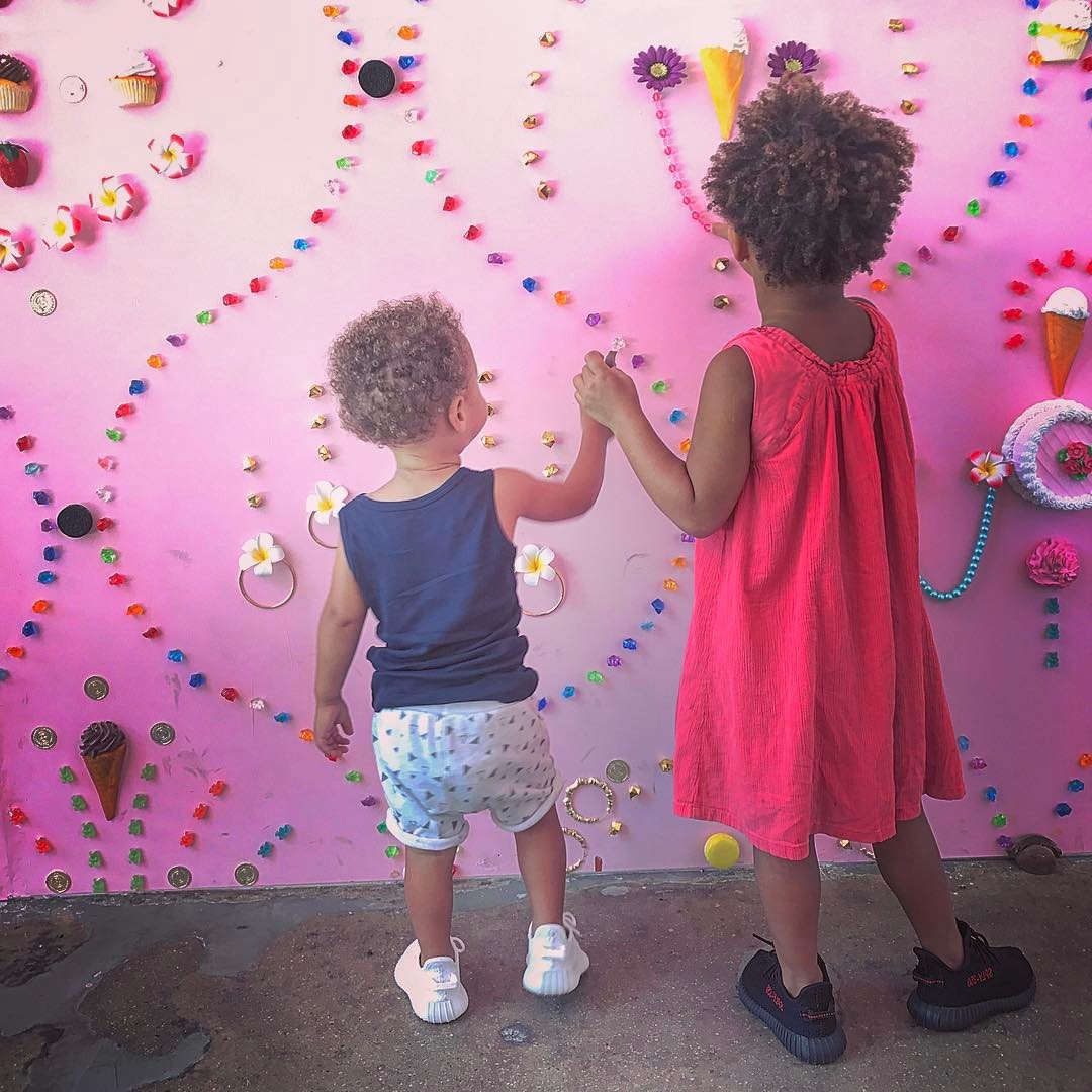 Two children, seen from behind, holding hands while looking at a pink wall adorned with colorful candy-like decorations.