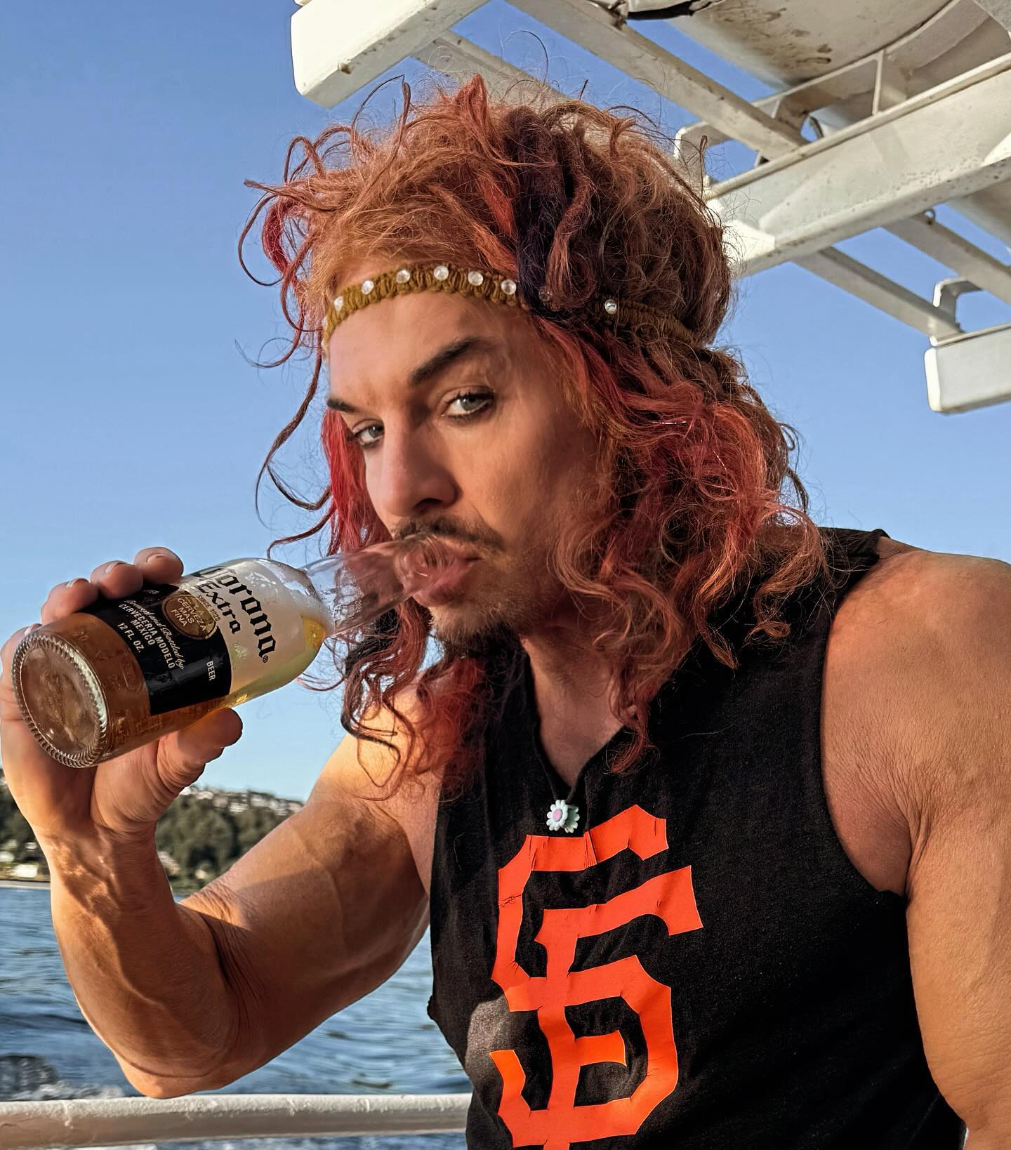 Carrot Top, wearing a black tank top and a beaded headband, drinks from a Corona bottle.