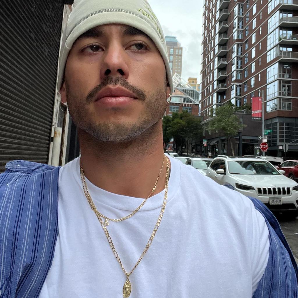 Ricky Alvarez in a white t-shirt and beanie, with a blue and white striped shirt draped over his shoulder, standing on a city street.