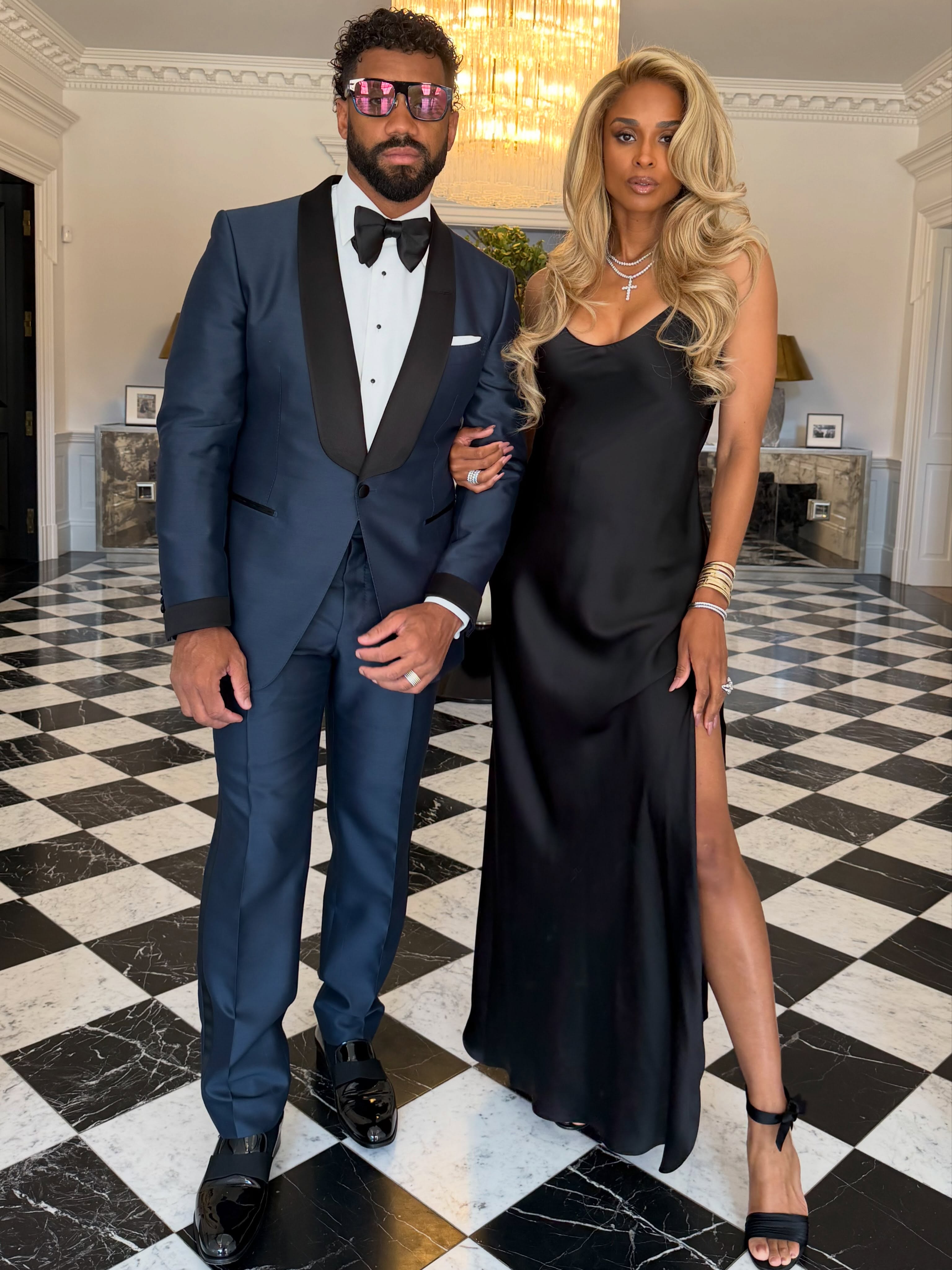 Russell Wilson and Ciara posing in formal attire on a black and white checkered floor.
